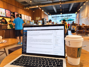 Laptop open to a WordPress blog editor beside a coffee cup in a busy café.