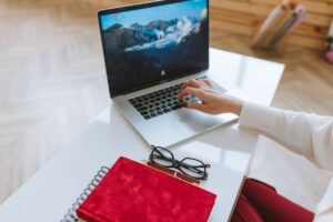 A person working on a laptop at a white desk with a red notebook, pen, and eyeglasses, representing remote work and freelancing.