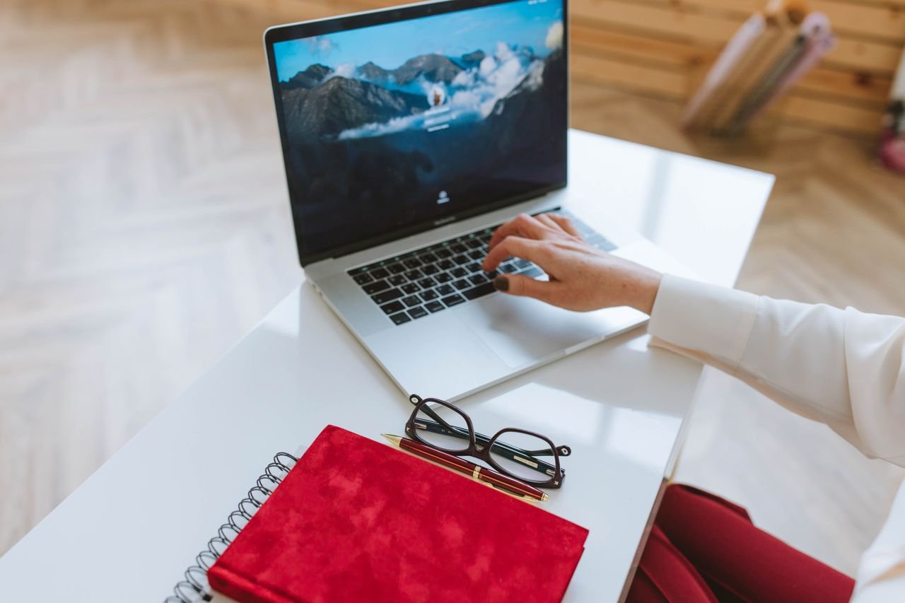 A person working on a laptop at a white desk with a red notebook, pen, and eyeglasses, representing remote work and freelancing.